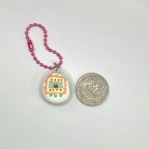 Puede incluir: Un juego electr&oacute;nico port&aacute;til blanco en forma de huevo, con un borde rosa y amarillo, sujeto a una cadena rosa. El juego tiene una peque&ntilde;a pantalla con caracteres japoneses. Una moneda de veinticinco centavos estadounidense est&aacute; al lado del juego para comparar el tama&ntilde;o.