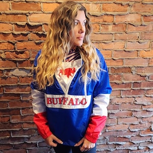 Può includere: Una giacca a vento blu, bianca e rossa con la scritta "BUFFALO" stampata sul davanti. La giacca ha una cerniera e un cappuccio. La persona che indossa la giacca è in piedi davanti a un muro di mattoni.