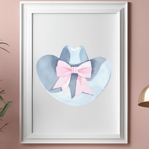 Op de afbeelding: Een ingelijste aquarelprint van een cowboyhoed met een roze strik. De hoed is lichtblauw en de strik is zachtroze. Het kunstwerk wordt weergegeven op een roze muur boven een roze kast.