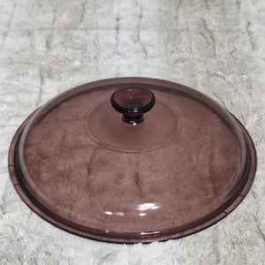 May include: A round, translucent purple glass lid featuring a central knob handle. The top surface of the lid displays a decorative radial sunburst pattern radiating from the center, with a smooth, slightly curved edge around the perimeter.
