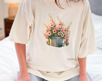 May include: A white oversized t-shirt with a floral print of pink flowers in a metal bucket. The text "Ivory" is printed on the bottom right corner of the shirt.