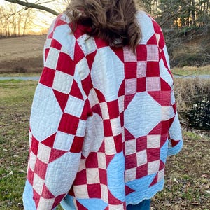 May include: A quilted jacket with a patchwork design in red, white, and light blue. The jacket features a variety of geometric shapes, including squares and diamonds. The edges of the jacket are trimmed with a light blue scalloped edge.