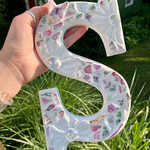 以下が含まれることがあります： 白いモザイク文字「S」は、花柄と蝶のアクセントで装飾されています。文字はピンク、緑、白の壊れた陶器の破片で装飾され、透明なガラスの装飾が施されています。文字は緑の草の背景の前に保持されています。