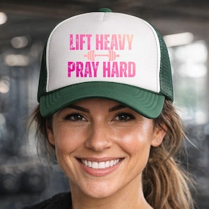 May include: A white and green trucker hat with the text "LIFT HEAVY PRAY HARD" in pink. The hat has a mesh back and a curved brim. The hat is worn by a person in a gym setting.