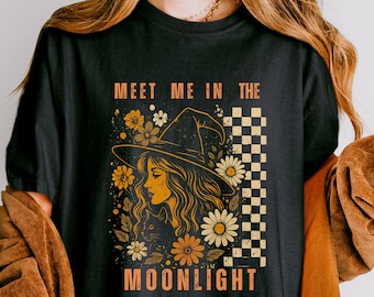 May include: A black t-shirt featuring a graphic print of a person wearing a wide-brimmed hat surrounded by flowers and a checkered pattern. The text "MEET ME IN THE MOONLIGHT" is printed in orange above and below the central illustration.