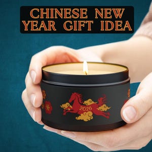 Op de afbeelding: Een brandende kaars in een zwarte blik met een gouden rand wordt in komvormige handen gehouden. De blik bevat een rood paardenontwerp met gouden wolken en het jaar 2026. De tekst "CHINESE NEW YEAR GIFT IDEA" staat bovenaan.