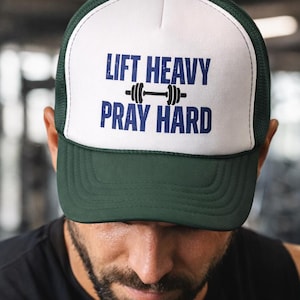 May include: A green and white trucker hat with the text "LIFT HEAVY PRAY HARD" in navy blue. The hat features a mesh back and a curved brim. The hat is worn by a person in a black tank top.