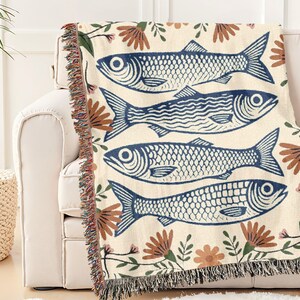 May include: A cream-colored throw blanket featuring a repeating pattern of blue fish illustrations. The blanket is adorned with floral accents in brown and pink, and has a fringed edge. The blanket is draped over a white sofa.