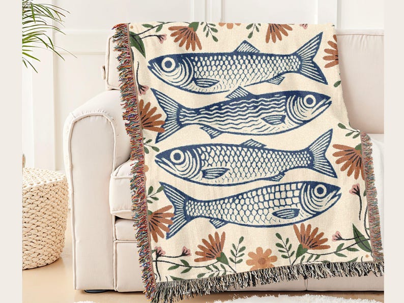 May include: A cream-colored throw blanket featuring a repeating pattern of blue fish illustrations. The blanket is adorned with floral accents in brown and pink, and has a fringed edge. The blanket is draped over a white sofa.