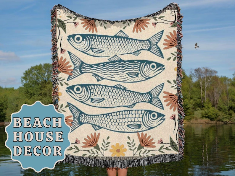 May include: Cream-colored woven throw blanket featuring four teal fish illustrations. The blanket has a floral and botanical design with a fringed edge. The words "BEACH HOUSE DECOR" are in a blue badge in the lower left corner.