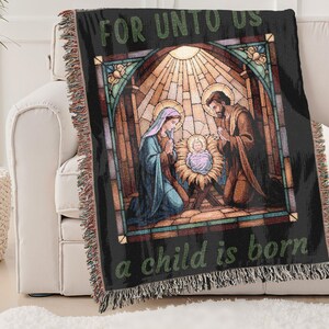 May include: A black woven throw blanket with a stained-glass style Nativity scene. The image shows Mary, Joseph, and baby Jesus. The text "FOR UNTO US a child is born" is printed on the blanket.