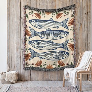 May include: Woven throw blanket with a cream background and a repeating pattern of four blue fish. The fish are surrounded by brown and green floral accents. The blanket has a fringed edge and is hanging on a wooden wall.