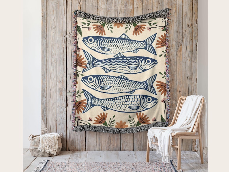 May include: Woven throw blanket with a cream background and a repeating pattern of four blue fish. The fish are surrounded by brown and green floral accents. The blanket has a fringed edge and is hanging on a wooden wall.