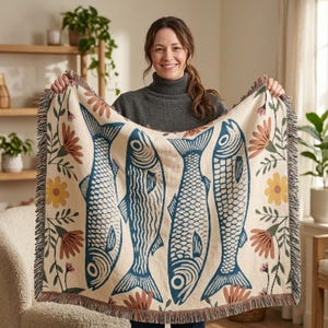 May include: A cream-colored throw blanket featuring four stylized blue fish designs. The blanket also includes floral and leaf patterns in shades of orange, yellow, and green. The edges are finished with a fringed trim, adding a decorative touch to the cozy home decor item.