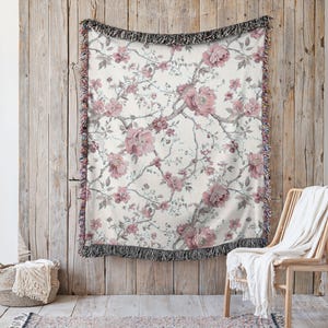 May include: A cream-coloured woven throw blanket featuring a floral pattern of pink peonies and grey branches. The blanket has a black fringe border and is displayed against a wooden wall. A wicker chair and basket are in the foreground.