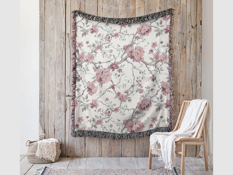 May include: A cream-coloured woven throw blanket featuring a floral pattern of pink peonies and grey branches. The blanket has a black fringe border and is displayed against a wooden wall. A wicker chair and basket are in the foreground.