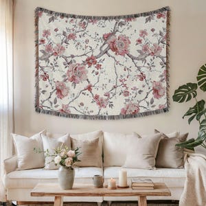 May include: Cream-coloured woven throw blanket with a floral pattern of pink peonies and grey branches. The blanket has a fringed edge and is displayed above a white sofa with beige cushions. Home decor.