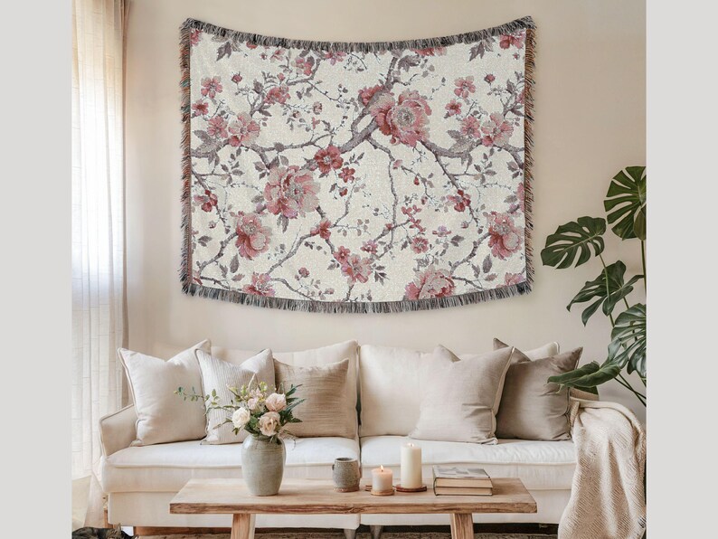 May include: Cream-coloured woven throw blanket with a floral pattern of pink peonies and grey branches. The blanket has a fringed edge and is displayed above a white sofa with beige cushions. Home decor.