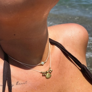 Può includere: Collana a due fili con un ciondolo a forma di conchiglia e piccole perline dorate. La collana è montata su un cordino bianco. Lo sfondo è una scena di spiaggia con il mare sullo sfondo. La collana è indossata sul collo di una persona.