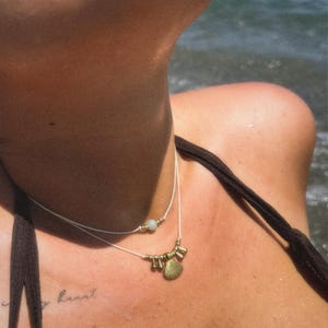 Può includere: Una collana a più strati con un ciondolo a forma di conchiglia e piccole perline color oro. La collana è indossata sul collo di una persona, con un tatuaggio visibile sulla pelle. Lo sfondo mostra l'oceano.