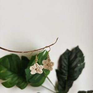 Op de afbeelding: Een paar crèmekleurige, gehaakte bloemoorbellen met goudkleurige haken en een kleine gouden kraal in het midden van elke bloem. De oorbellen hangen aan een dunne, bruine tak, met groene bladeren op de achtergrond.