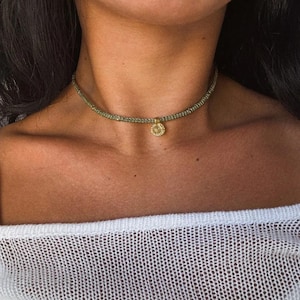 Peut inclure: Un collier ras du cou vert avec un petit pendentif doré. Le pendentif présente un motif de soleil. Le collier est porté autour du cou.