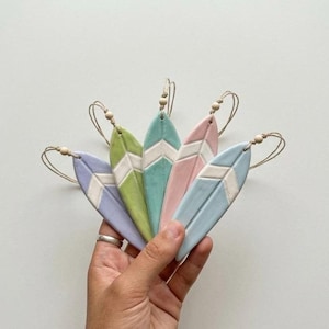 Peut inclure: Cinq ornements en céramique en forme de planche de surf dans des tons pastel de lavande, vert lime, menthe, rose et bleu clair. Chaque pièce présente un motif chevron blanc central et est fixée à une boucle en ficelle avec une perle en bois.