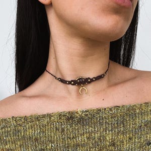 May include: A dark brown macrame choker necklace with gold-colored beads and a gold crescent moon pendant. The necklace is worn around the neck. The person is wearing a green sweater.