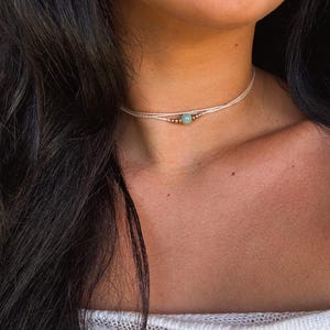 Può includere: Una delicata collana girocollo con una gemma verde chiaro e piccole perline dorate. La collana è infilata su un cordoncino color crema. Il gioiello è indossato intorno al collo.