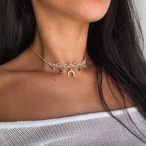 May include: A delicate macrame choker necklace with a gold crescent moon pendant. The necklace features small, round beads and dangling accents. The choker is worn around the neck, showcasing the intricate design and craftsmanship.