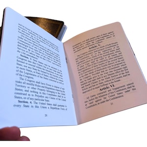 May include: Open book with text from the United States Constitution. The pages are white with black text, and the book has a dark brown cover. The text includes articles and sections, with page numbers visible at the bottom.