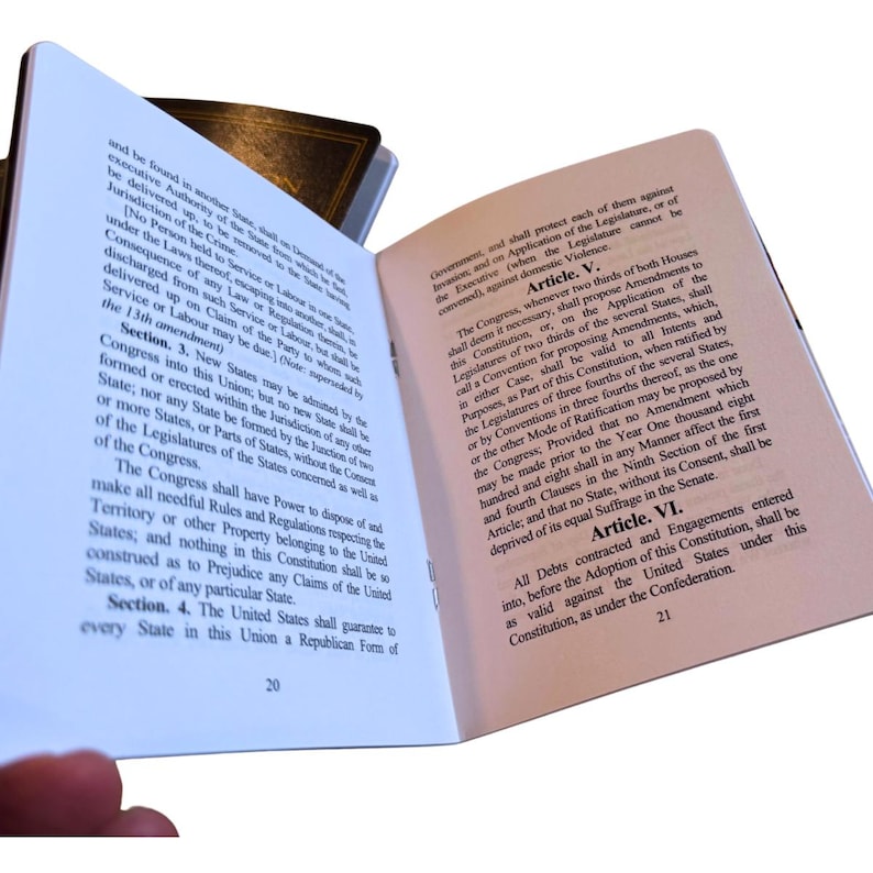 May include: Open book with text from the United States Constitution. The pages are white with black text, and the book has a dark brown cover. The text includes articles and sections, with page numbers visible at the bottom.
