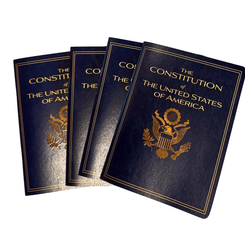 May include: Four navy blue books titled "The Constitution of The United States of America" with gold lettering and a gold emblem. The books are slightly fanned out, with the front cover of the top book visible.