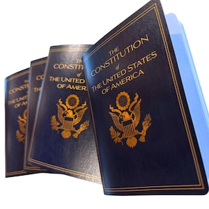 May include: A stack of dark blue books with gold lettering and a gold emblem. The text on the cover reads "THE CONSTITUTION OF THE UNITED STATES OF AMERICA". The books are slightly angled, revealing the pages.