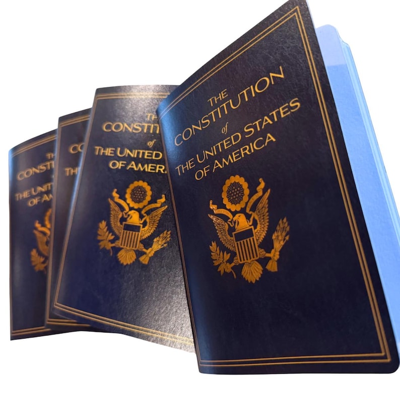 May include: A stack of dark blue books with gold lettering and a gold emblem. The text on the cover reads "THE CONSTITUTION OF THE UNITED STATES OF AMERICA". The books are slightly angled, revealing the pages.
