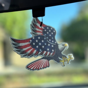 May include: A red, white and blue eagle air freshener hanging from a rear-view mirror in a car. The eagle is flying with its wings spread and has a star-spangled design.