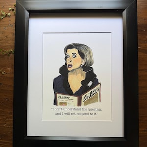 Può includere: Stampa artistica incorniciata con un'illustrazione ad acquerello di una donna sorpresa, che tiene in mano una rivista. La rivista recita "KLIMPY'S" e la citazione "Non capisco la domanda e non risponderò". L'opera d'arte è in una cornice nera.