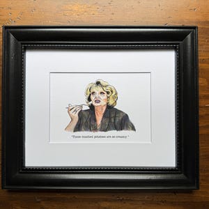 Könnte beinhalten: Gerahmter Aquarelldruck einer Frau mit blonden Haaren, die eine Gabel hält. Der Druck befindet sich in einem schwarzen Rahmen mit weißem Passepartout. Der Text lautet: "These mashed potatoes are so creamy."