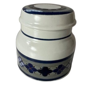 May include: A ceramic jar with a lid. The jar is primarily off-white with a blue and gray floral pattern around the base. The lid has a dark blue rim and a white center with small dark blue circles.