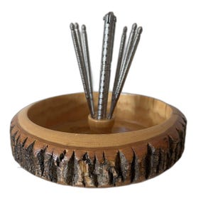May include: A wooden nut bowl with a natural bark edge and a set of silver nutcrackers. The bowl is light brown with a dark brown bark edge. The nutcrackers are arranged in the center of the bowl.