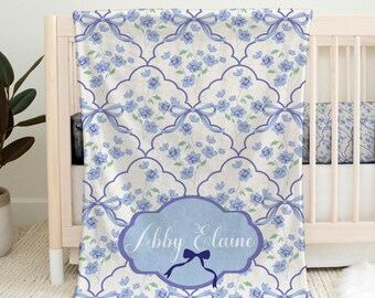 Puede incluir: Una manta rectangular para bebé con fondo blanco, un patrón floral azul repetitivo, lazos azules y una etiqueta central azul festoneada que contiene el texto Libby Elaine en letra cursiva blanca con un pequeño icono de lazo azul debajo.