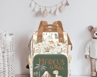 May include: A green and brown diaper bag with a dinosaur print. The bag has a large pocket on the front with the name "Marcus White" printed in white.