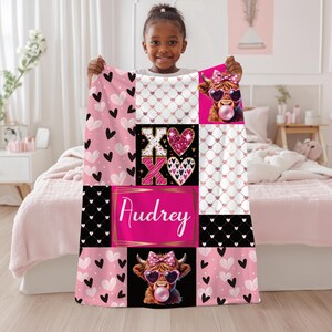 May include: A rectangular fleece blanket featuring a patchwork design with pink and black hearts, a glittery XOXO graphic, and two illustrations of a highland cow wearing heart-shaped sunglasses and blowing a bubble. The name Audrey is printed in white.