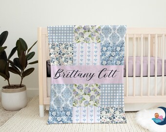 Puede incluir: Una manta para bebé con un diseño de patchwork en tonos azules, blancos y lavanda, con estampados florales y de cuadros. El nombre "Brittany Cott" se muestra en una pancarta rosa. La manta está sobre una cuna.