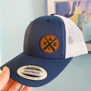 Puede incluir: Una gorra de béisbol azul marino y blanca con un parche de cuero marrón que dice "DAD" con bates de béisbol cruzados. El parche tiene un borde cosido. La gorra tiene una parte posterior de malla y una etiqueta que dice "The Authentic Yupoong Snapback Yupoong Since 1974".