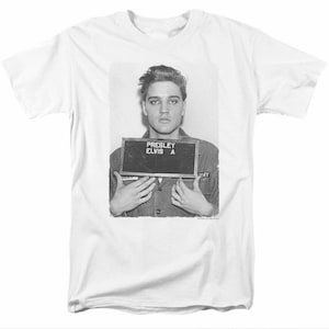 May include: Black and white photo of Elvis Presley in a military uniform, holding a sign that reads "PRESLEY ELVIS A".