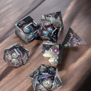 May include: A set of seven iridescent dice with a black and blue galaxy-like swirl pattern. The dice are made of clear resin and have silver numbers. Each die has a pearl-like sphere embedded in the resin.