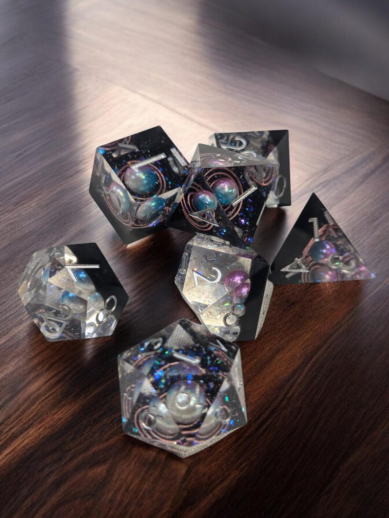May include: A set of six clear resin dice with iridescent glitter and embedded pearls. The dice are numbered 1 through 6 and have a galaxy-themed design.