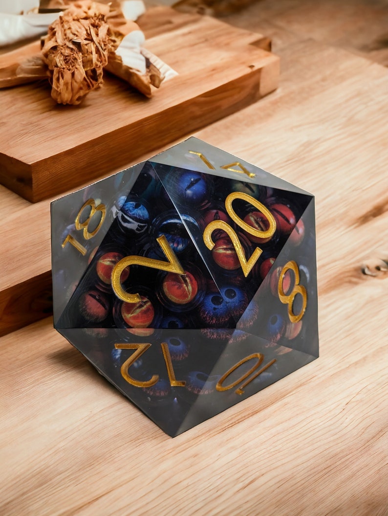 May include: A black and gold 20-sided die with a design of eyes and a dark background. The die is made of clear plastic and has gold numbers.