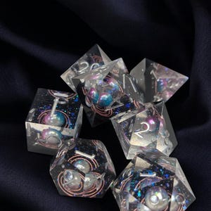 May include: A set of seven clear resin dice with iridescent glitter and embedded pearls. The dice are in various shapes and sizes, with numbers visible on each face.